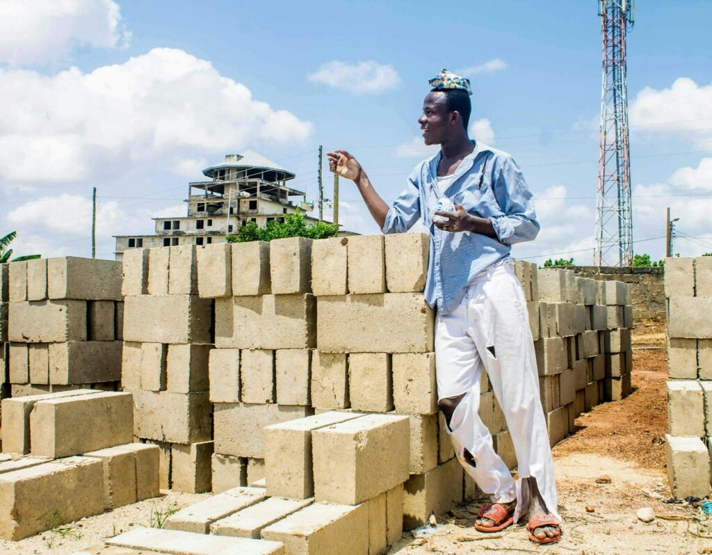 A young man stands in a construction site surrounded by concrete blocks, with a building and tower in the background.