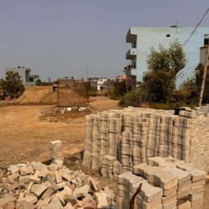 Image of a rural construction site featuring organized stacks of bricks and distant residential buildings.
